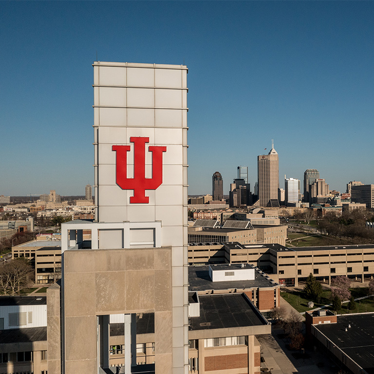 Photo of the top of the Bell Tower of the Campus Center that features a crimson IU trident at the top. The Indianapolis skyline is in the background.